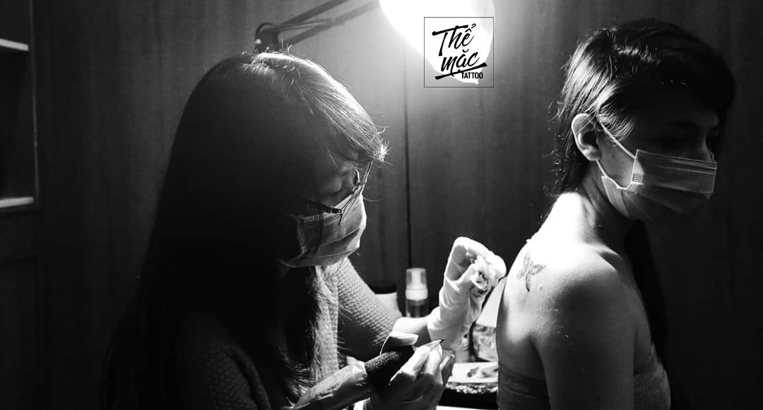A black and white photo of a tattoo artist working on a client's upper back — both wearing face masks, captured in an intimate and focused moment.