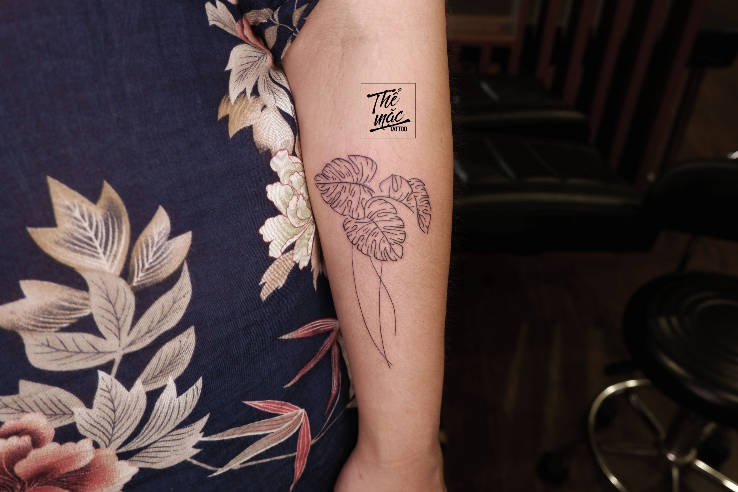 A fine line tattoo of large tropical monstero leaves with long stems, placed on the forearm — organic and flowing in style.
