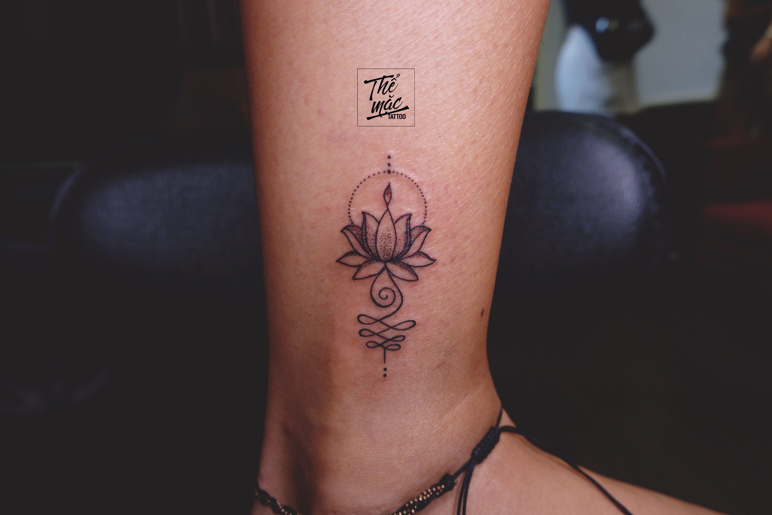 A spiritual fine line tattoo featuring a lotus flower, unalome symbol, and dotted halo — inked on the ankle.