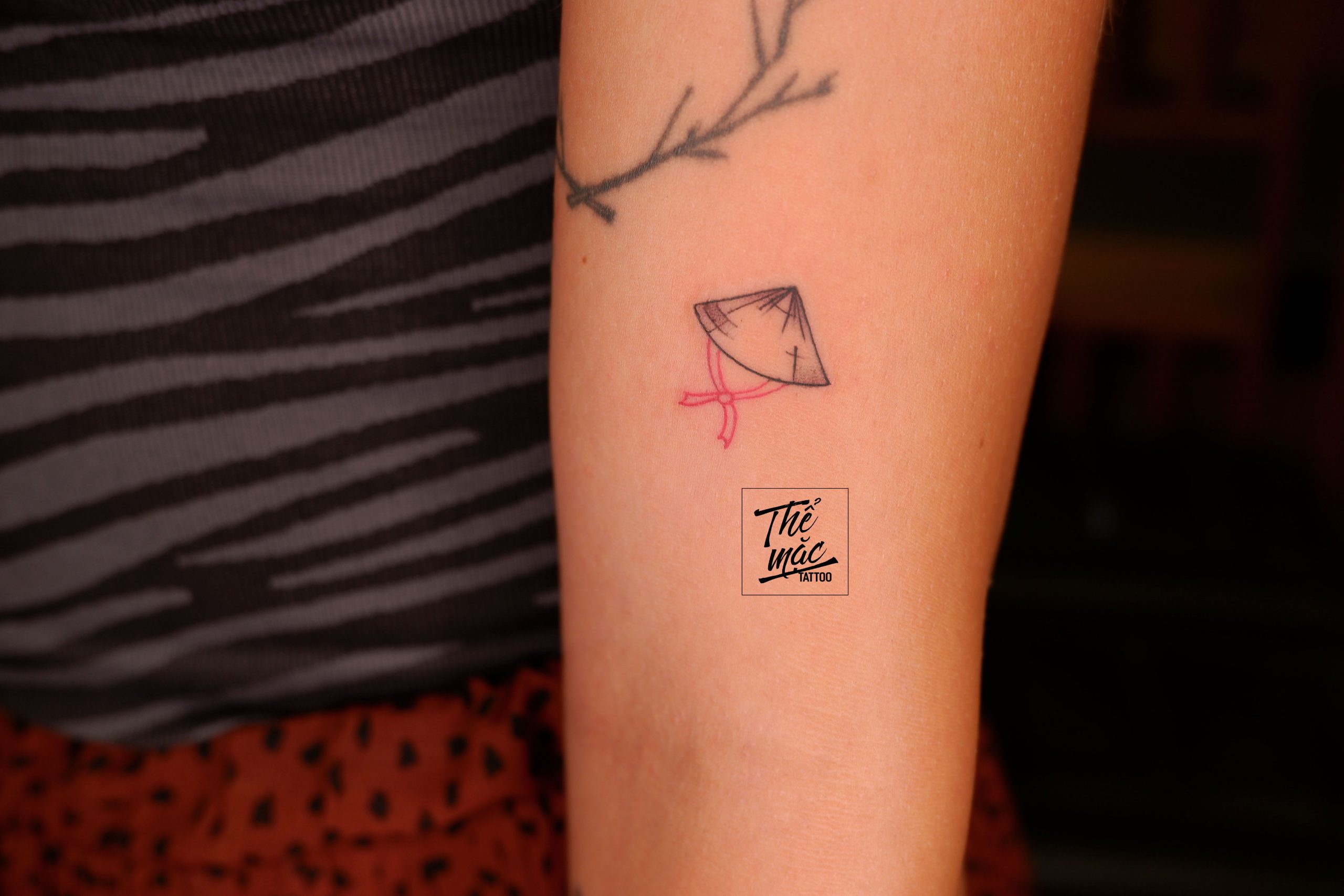 A minimalist fine line tattoo of a Vietnamese conical hat (nón lá) with a pink ribbon, placed on the inner arm — a meaningful symbol of Vietnamese culture.