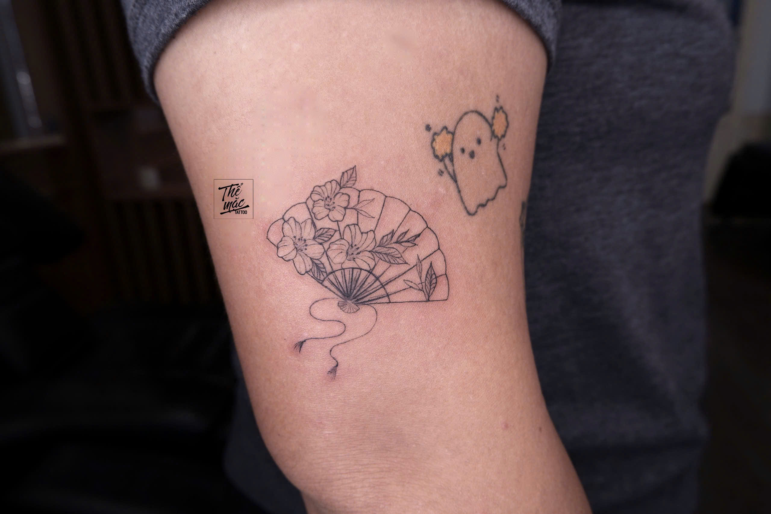 A detailed fine line tattoo of a traditional hand fan filled with blooming flowers and leaves — inked on the upper arm.